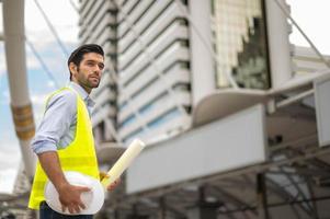 Kaukasisch Mens ingenieur , vervelend geel hesje en groot moeilijk hoed, en hand- Holding de wit verdieping plan in de plaats werk van de centrum stad. foto