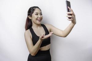 een portret van een sportief Aziatisch vrouw vervelend een sportkleding en Holding haar telefoon, geïsoleerd door wit achtergrond foto