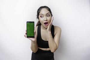 geschokt sportief Aziatisch vrouw vervelend sportkleding en tonen groen scherm Aan haar telefoon, geïsoleerd door wit achtergrond foto
