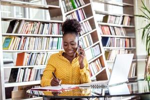 geslaagd bedrijf vrouw werken Bij de kantoor, Afrikaanse Amerikaans vrouw pratend Aan haar mobiel telefoon en werken Aan laptop. klein bedrijf ondernemer concept foto