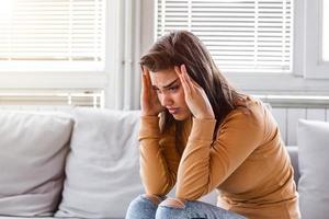 portret van een jong brunette meisje zittend Aan de bankstel Bij huis met een hoofdpijn en migraine . mooi vrouw lijden van chronisch dagelijks hoofdpijn. verdrietig vrouw Holding haar hoofd omdat sinus pijn foto