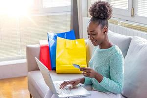 jong vrouw Aan sofa boodschappen doen online met laptop. jong zwart vrouw invoeren kaart informatie terwijl boodschappen doen online. online boodschappen doen concept foto