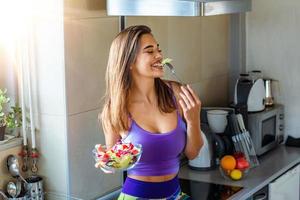 jong en gelukkig vrouw aan het eten gezond salade zittend Aan de keuken bureau met groen vers ingrediënten binnenshuis. mooi fit vrouw aan het eten gezond salade na geschiktheid training foto