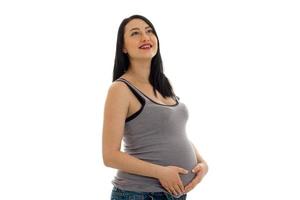 studio portret van jong zwanger brunette vrouw in overhemd aanraken haar buik en op zoek omhoog en glimlachen geïsoleerd Aan wit achtergrond foto