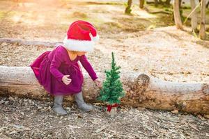 schattig gemengd ras jong baby meisje hebben pret met de kerstman hoed en Kerstmis boom buitenshuis Aan log foto