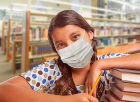 spaans meisje leerling vervelend gezicht masker aan het studeren in bibliotheek foto