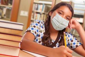 spaans meisje leerling vervelend gezicht masker aan het studeren in bibliotheek foto