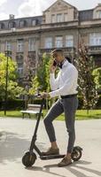 jonge Afro-Amerikaan die mobiele telefoon gebruikt terwijl hij met een elektrische scooter op straat staat foto