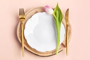 tafel instelling - gouden bestek en een roze tulp Aan wit bord Aan houten zag besnoeiing Aan pastel roze. foto
