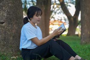 Aziatisch vrouw zittend met haar terug tegen een boom lezing een boek. concept. Aziatisch vrouw aan het doen buitenshuis activiteiten, zo net zo lezing boeken, werken, hebben een picknick met familie.zacht en selectief focus . foto