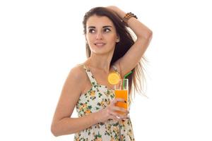 jong mooi brunette meisje in sarafan met bloemen patroon drankjes oranje cocktail en looks terzijde geïsoleerd Aan wit achtergrond foto