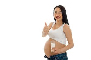 portret van jong grappig zwanger vrouw met glas van melk tonen duimen omhoog Aan camera geïsoleerd Aan wit achtergrond foto