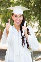 gemengd ras duimen omhoog meisje vieren diploma uitreiking buiten in pet en japon met diploma in hand- foto