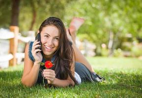 aantrekkelijk gelukkig gemengd ras jong vrouw pratend Aan cel telefoon buiten houdende in de gras foto