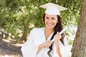 aantrekkelijk gemengd ras meisje vieren diploma uitreiking buiten in pet en japon met diploma in hand- foto