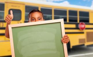 jong spaans jongen met blanco schoolbord in de buurt school- bus foto