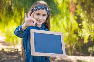 schattig jong gemengd ras meisje met Oke teken Holding blanco schoolbord buitenshuis foto