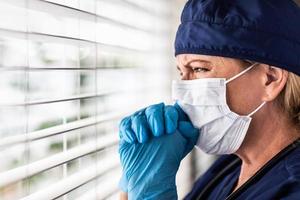 biddend benadrukt vrouw dokter of verpleegster Aan breken Bij venster vervelend medisch gezicht masker foto