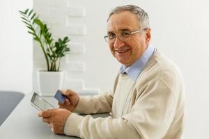 knap vrolijk senior Mens gebruik makend van digitaal tablet zittend Bij de tafel in de leven kamer. vrije tijd activiteiten, uitgeven tijd, gelukkig pensioen en senior levensstijl concept foto