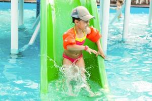 gelukkig weinig meisje Speel Aan water glijbaan in buitenshuis zwemmen zwembad Aan heet zomer dag. kinderen leren naar zwemmen. kinderen vervelend zon bescherming uitslag bewaker glijden Aan aqua speelplaats in tropisch toevlucht foto