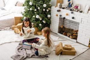 twee meisjes in voorkant van Kerstmis boom met cadeaus en brand plaats foto