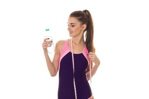 schattig slank jong brunette meisje in lichaam zwempak met meten plakband Aan haar nek en fles van water in handen geïsoleerd Aan wit achtergrond foto
