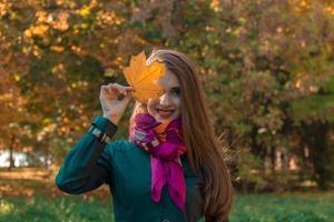 mooi meisje in sjaal houdt herfst blad in de buurt de ogen en glimlacht foto