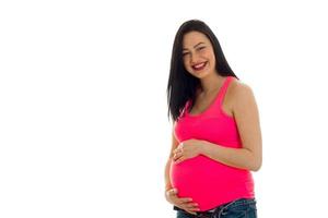 vrolijk jong brunette zwanger meisje in roze overhemd met hand- Aan haar buik poseren geïsoleerd Aan wit achtergrond foto