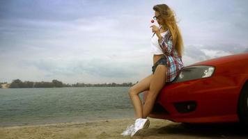 sensueel blond in bril zit Aan een rood auto met lolly in haar handen foto