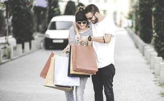 portret van vrolijk Kaukasisch jong paar Mens en vrouw Holding veel papier Tassen na boodschappen doen terwijl wandelen en pratend Aan straat. gelukkig familie paar met pakketjes buitenshuis. buying concept foto