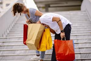 portret van vrolijk Kaukasisch jong paar Mens en vrouw Holding veel papier Tassen na boodschappen doen terwijl wandelen en pratend Aan straat. gelukkig familie paar met pakketjes buitenshuis. buying concept foto