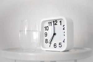 wit alarm klok en glas van water Aan tafel in slaapkamer. wakker worden omhoog vroeg in de ochtend- door de alarm klok en drinken water. foto