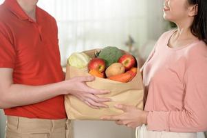 Aziatisch jong Mens levering in rood uniform Holding gezond fruit groenten in kruidenier zak sturen naar klant in keuken achtergrond foto