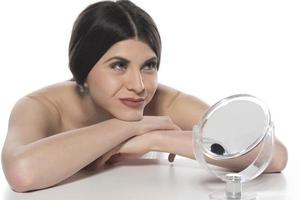 mooi aantrekkelijk brunette vrouw controle haar gezicht huid in voorkant van een spiegel Aan wit achtergrond. foto