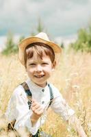 weinig boer Aan een zomer veld, schattig weinig jongen in een rietje hoed. jongen in een hoed met een bloem staat in een veld. portret van een dorp jongen foto