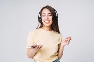 emotioneel portret van jong gelukkig Aziatisch vrouw met lang haar- in geel overhemd en jeans gebruik makend van mobiel telefoon luister muziek- door hoofdtelefoons Aan grijs achtergrond foto