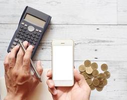 hand- met zilver pen gebruik makend van rekenmachine en Holding smartphone wit scherm met goud munten groep Aan wit hout tafel top visie en kopiëren ruimte natuur schaduw foto