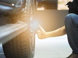 Mens hand- duim omhoog in de buurt groot wiel van nieuw auto met licht gloed foto