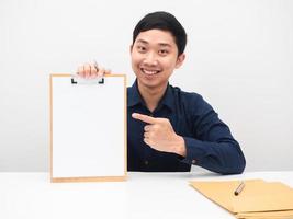 knap Mens glimlach gezicht zittend Bij de tafel en punt vinger Bij blanco papier Aan houten bord wit achtergrond foto