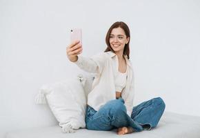 mooi glimlachen vrouw tiener meisje leerling met donker lang haar- in wit overhemd nemen selfie Aan mobiel telefoon in hand- zittend Aan bankstel Bij huis foto