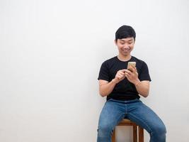 Aziatisch Mens gebruik makend van smartphone in hand- zittend Aan stoel gelukkig glimlach gezicht Aan wit muur achtergrond