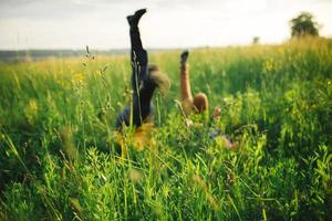 vrouw en Mens hebben pret buitenshuis. liefhebbend hipster paar wandelen in de veld, zoenen en Holding handen, knuffelen, aan het liegen in de gras en hijs- hun poten omhoog in de zomer Bij zonsondergang. valentijnsdag dag foto