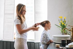 jong moeder vrouw met lang haar- met weinig tween meisje dochter in pyjama kammen haar haar- Aan ochtend- in badkamer Bij huis foto