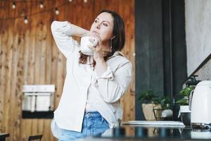 volwassen midden- oud brunette glimlachen vrouw vijftig jaren in wit overhemd met kop van ochtend- koffie in keuken Bij huis foto