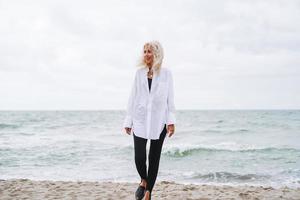 portret van elegant blond vrouw in wit overhemd Aan zand strand Bij storm zee Bij winderig weater foto