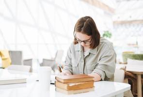 jong brunette tiener meisje college leerling in bril aan het doen huiswerk met boeken Bij groen modern bibliotheek openbaar plaats foto