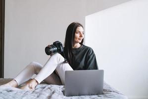 jong vrouw fotograaf met digitaal camera freelancer in gewoontjes kleren gebruik makend van laptop zittend Aan bed Bij huis foto