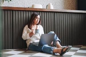 volwassen brunette midden- oud vrouw vijftig jaren freelancer in gewoontjes kleren met ochtend- koffie gebruik makend van laptop zittend Aan bankstel Bij de huis foto