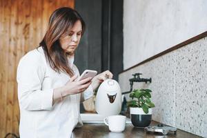 volwassen midden- oud brunette glimlachen vrouw vijftig jaren in wit overhemd gebruik makend van mobiel telefoon met kop van ochtend- koffie in keuken Bij huis foto