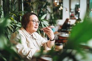 volwassen brunette midden- oud vrouw plus grootte lichaam positief in gewoontjes kleren met papier kop van koffie gebruik makend van mobiel telefoon in cafe foto